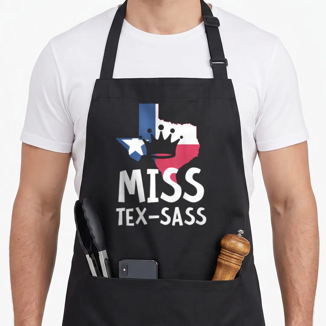 Miss texans apron - Grey crown on Texas map in blue white red with Miss Tex-Sass text — shown on a black cotton canvas apron against a white background