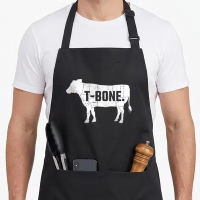 Beef steak apron - White distressed cow silhouette with T-BONE text and dashed butcher cut lines — shown on a black cotton canvas apron against a white background