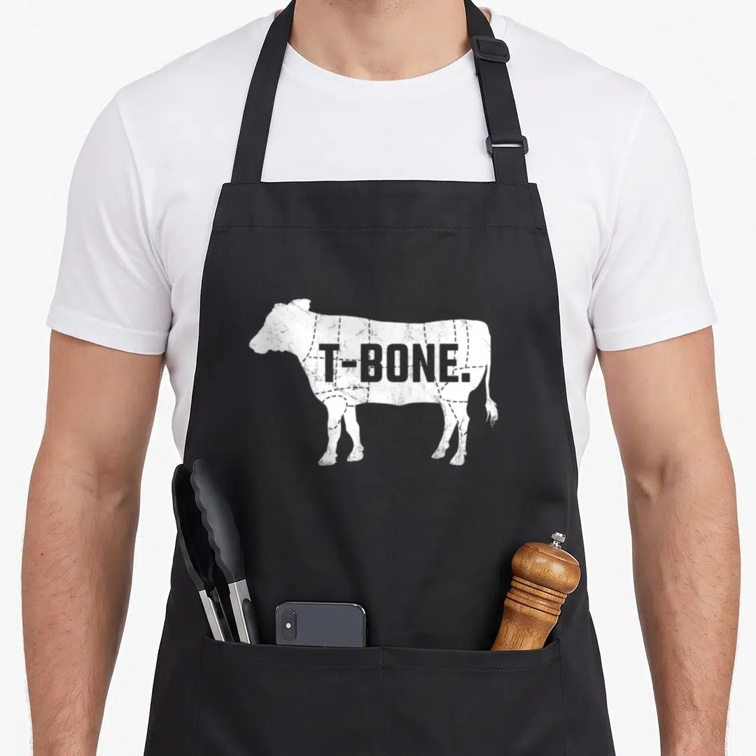 Beef steak apron - White distressed cow silhouette with T-BONE text and dashed butcher cut lines — shown on a black cotton canvas apron against a white background