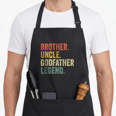 Uncle goddaughter apron - Stacked text BROTHER UNCLE GODFATHER LEGEND in orange yellow beige green retro font — shown on a black cotton canvas apron against a white background