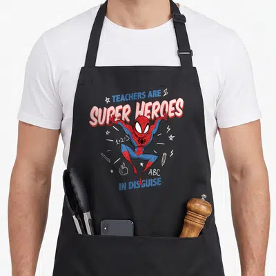 Cute heroe teacher apron - Spider-Man in red blue white with TEACHERS ARE SUPER HEROES IN DISGUISE text and school — shown on a black cotton canvas apron against a white background
