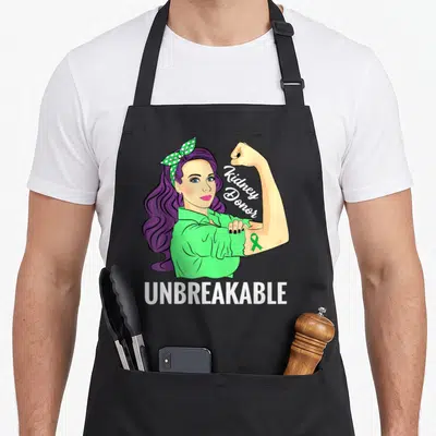 Kidney transplant awareness apron - Cartoon woman flexing arm with green ribbon and Kidney Donor UNBREAKABLE text — shown on a black cotton canvas apron against a white background