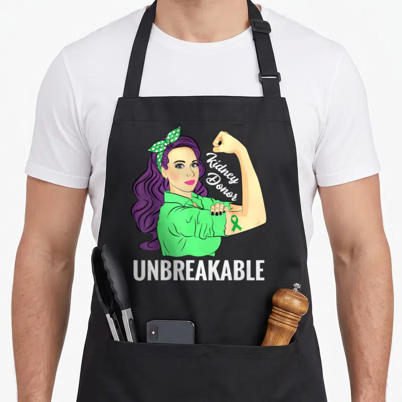 Kidney transplant awareness apron - Cartoon woman flexing arm with green ribbon and Kidney Donor UNBREAKABLE text — shown on a black cotton canvas apron against a white background
