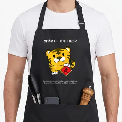 Tiger chinese zodiac aprons - Cartoon tiger holding red sign with character and list of personality traits — shown on a black cotton canvas apron against a white background
