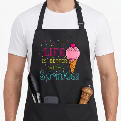 Better sprinkle apron - Cartoon pink ice cream cone with LIFE IS BETTER WITH Sprinkles text and scattered sprinkles — shown on a black cotton canvas apron against a white background