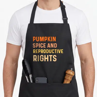 Reproductive spice apron - Stacked orange peach and yellow text reading PUMPKIN SPICE AND REPRODUCTIVE RIGHTS — shown on a black cotton canvas apron against a white background