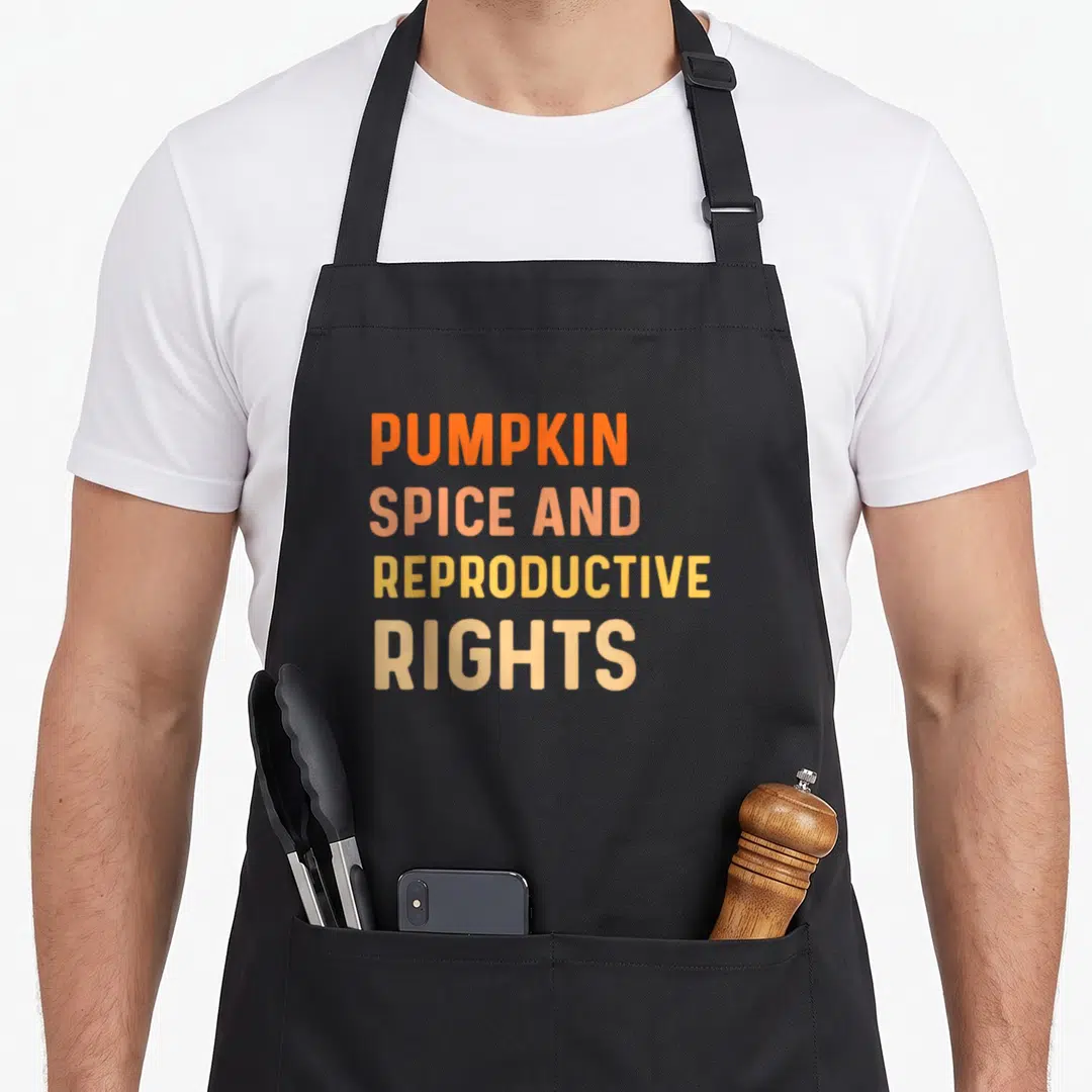 Reproductive spice apron - Stacked orange peach and yellow text reading PUMPKIN SPICE AND REPRODUCTIVE RIGHTS — shown on a black cotton canvas apron against a white background