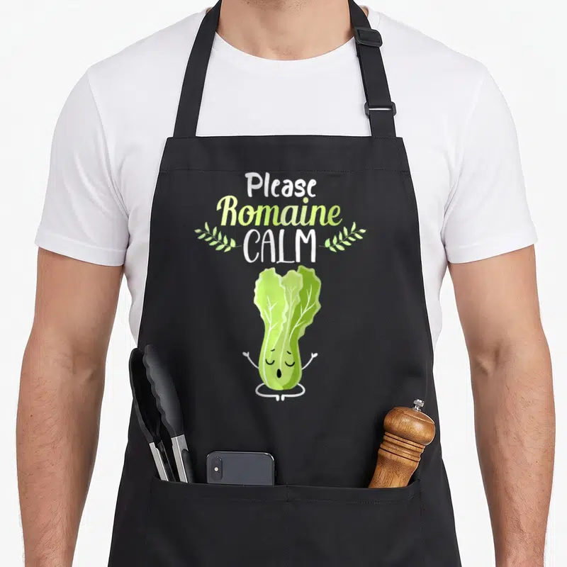 Romaine vegetable apron - Please Romaine Calm text with meditating cartoon romaine lettuce and two leaf sprigs — shown on a black cotton canvas apron against a white background