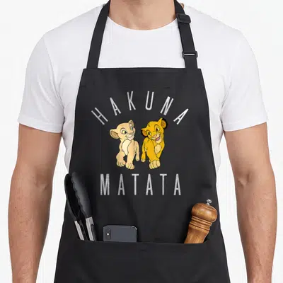Simba nala lion apron - Two lion cubs under arched HAKUNA MATATA text in yellow tan white cartoon style — shown on a black cotton canvas apron against a white background