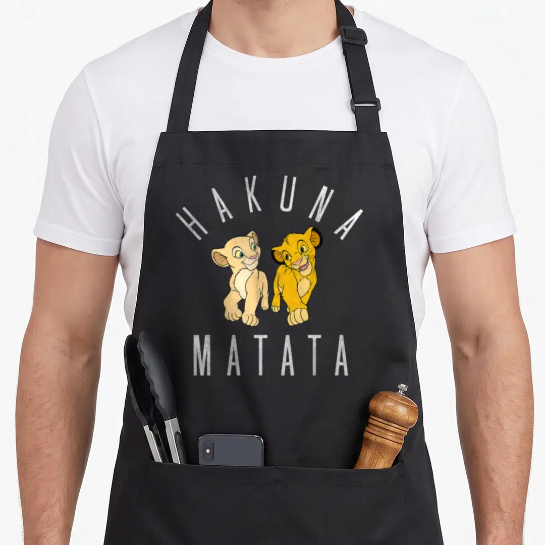 Simba nala lion apron - Two lion cubs under arched HAKUNA MATATA text in yellow tan white cartoon style — shown on a black cotton canvas apron against a white background