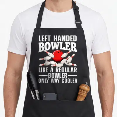 Handed left apron - Red bowling ball hitting white pins with white text on grey background — shown on a black cotton canvas apron against a white background