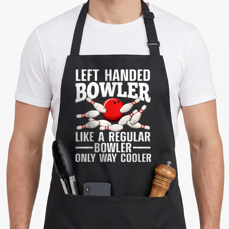 Handed left apron - Red bowling ball hitting white pins with white text on grey background — shown on a black cotton canvas apron against a white background
