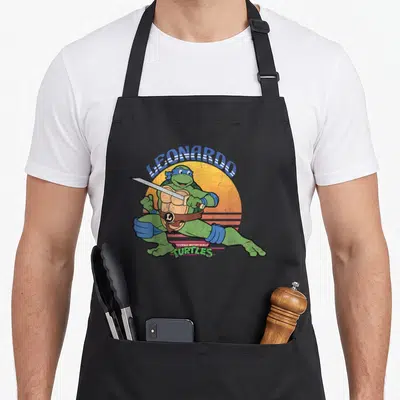 Leonardo sun teenage cooking aprons - Leonardo leaping with katanas over orange sun with blue and pink lettering — shown on a black cotton canvas apron against a white background