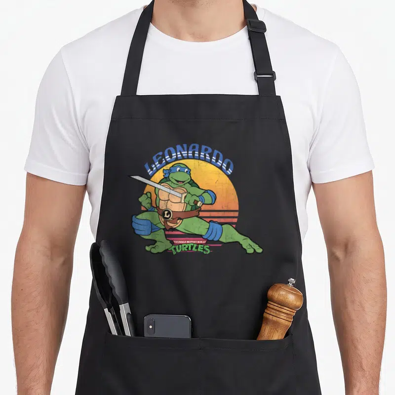 Leonardo sun teenage cooking aprons - Leonardo leaping with katanas over orange sun with blue and pink lettering — shown on a black cotton canvas apron against a white background