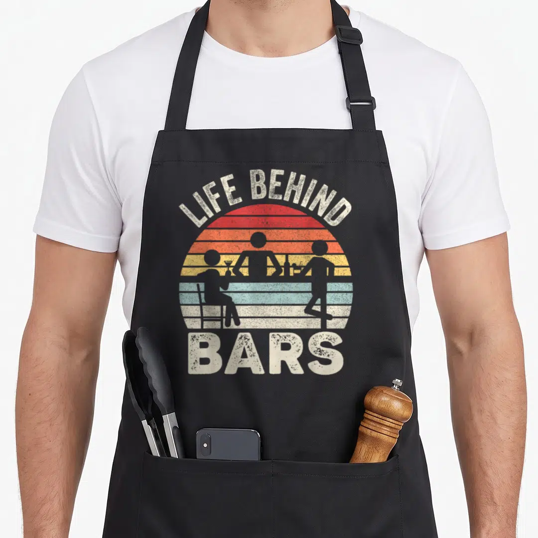 Behind bartender apron - Retro striped sun with silhouettes of people at a bar and arched text reading Life Behind Bars — shown on a black cotton canvas apron against a white background