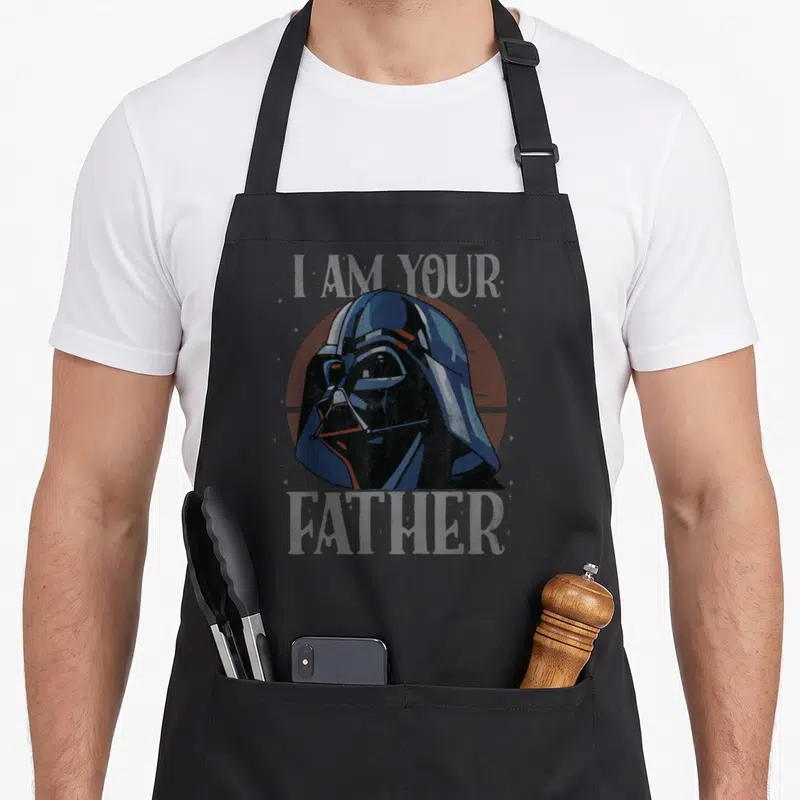 Cute father falcon dad apron - Darth Vader head over red circle with I AM YOUR FATHER text in distressed blue and grey — shown on a black cotton canvas apron against a white background