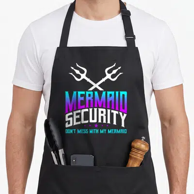 Merdad grill daddy apron - Crossed white tridents above gradient text reading MERMAID SECURITY DON'T MESS WITH MY — shown on a black cotton canvas apron against a white background