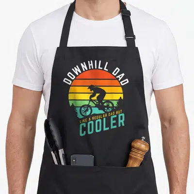 Downhill biking dad apron - Retro striped sun with black cyclist silhouette and text Downhill Dad Like A Regular Dad — shown on a black cotton canvas apron against a white background