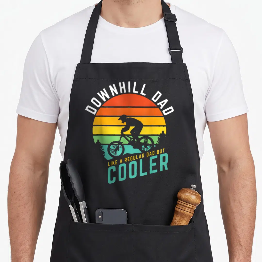 Downhill biking dad apron - Retro striped sun with black cyclist silhouette and text Downhill Dad Like A Regular Dad — shown on a black cotton canvas apron against a white background
