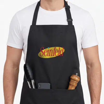Original colorful apron - Red Seinfeld script text on yellow oval with small blue triangle above — shown on a black cotton canvas apron against a white background