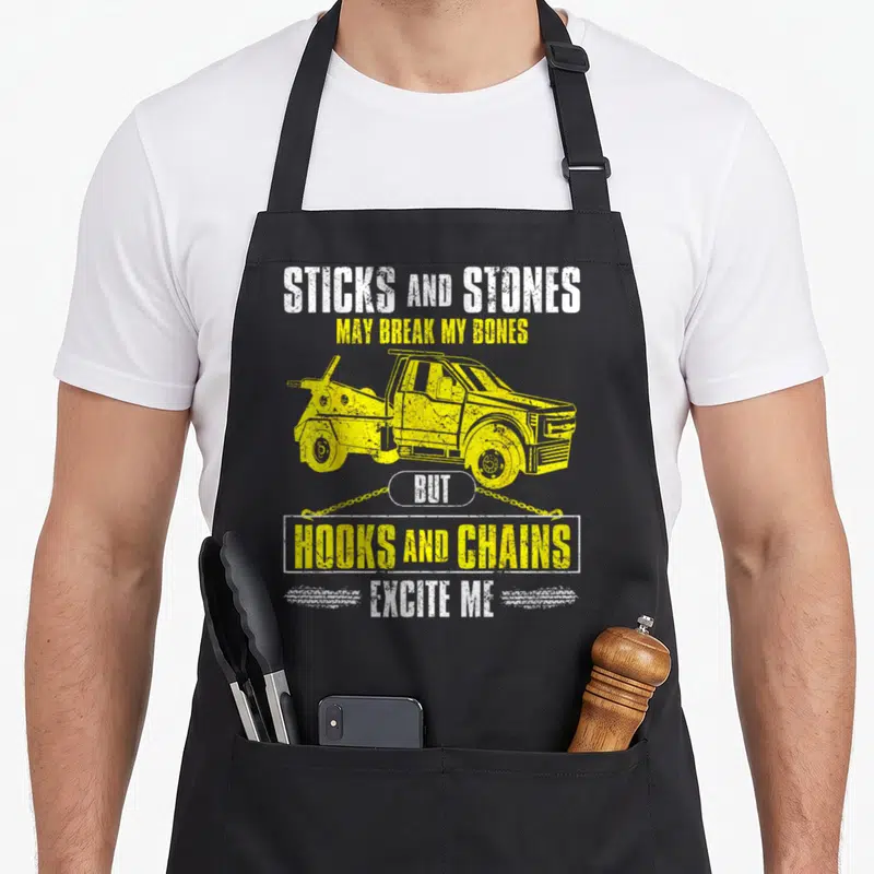 Breakdown truck apron - Yellow tow truck graphic with stacked text Sticks and Stones May Break My Bones But Hooks — shown on a black cotton canvas apron against a white background