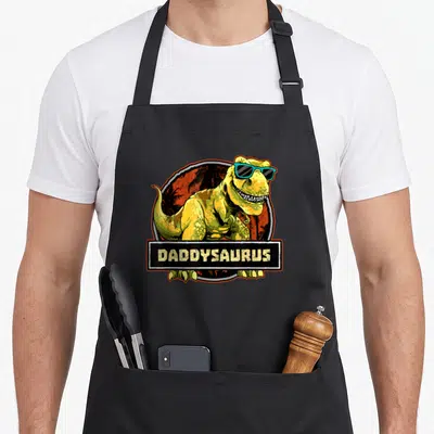 Daddysaurus dinosaur father day apron - Yellow T-Rex wearing blue sunglasses inside orange circle with black banner — shown on a black cotton canvas apron against a white background