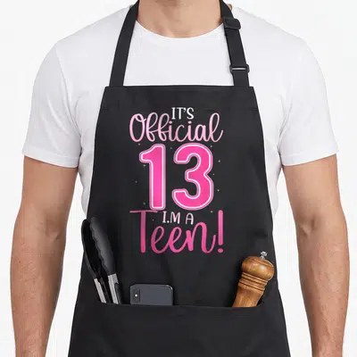 13th age birthday apron - Pink and white text IT'S Official 13 I'M A Teen! with sparkles — shown on a black cotton canvas apron against a white background