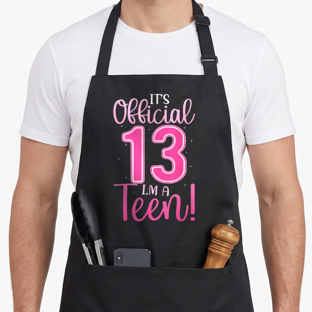 13th age birthday apron - Pink and white text IT'S Official 13 I'M A Teen! with sparkles — shown on a black cotton canvas apron against a white background