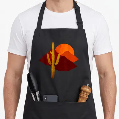 Saguaro sunset cactus apron - Minimalist yellow cactus standing before red mountains and orange sun — shown on a black cotton canvas apron against a white background