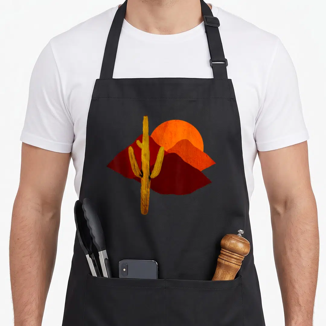 Saguaro sunset cactus apron - Minimalist yellow cactus standing before red mountains and orange sun — shown on a black cotton canvas apron against a white background