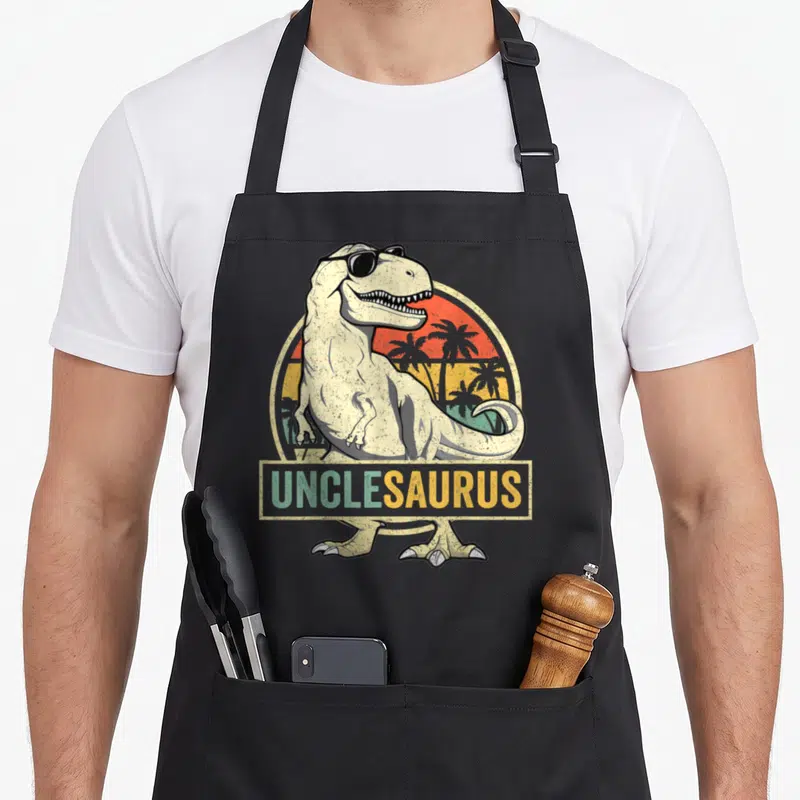 Uncle dinosaur apron - Cartoon T-Rex in sunglasses with UNCLESAURUS text over retro sunset — shown on a black cotton canvas apron against a white background