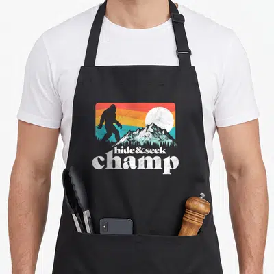 Champ bigfoot apron - Distressed hide and seek champ text below Bigfoot silhouette against colorful mountain sunset — shown on a black cotton canvas apron against a white background