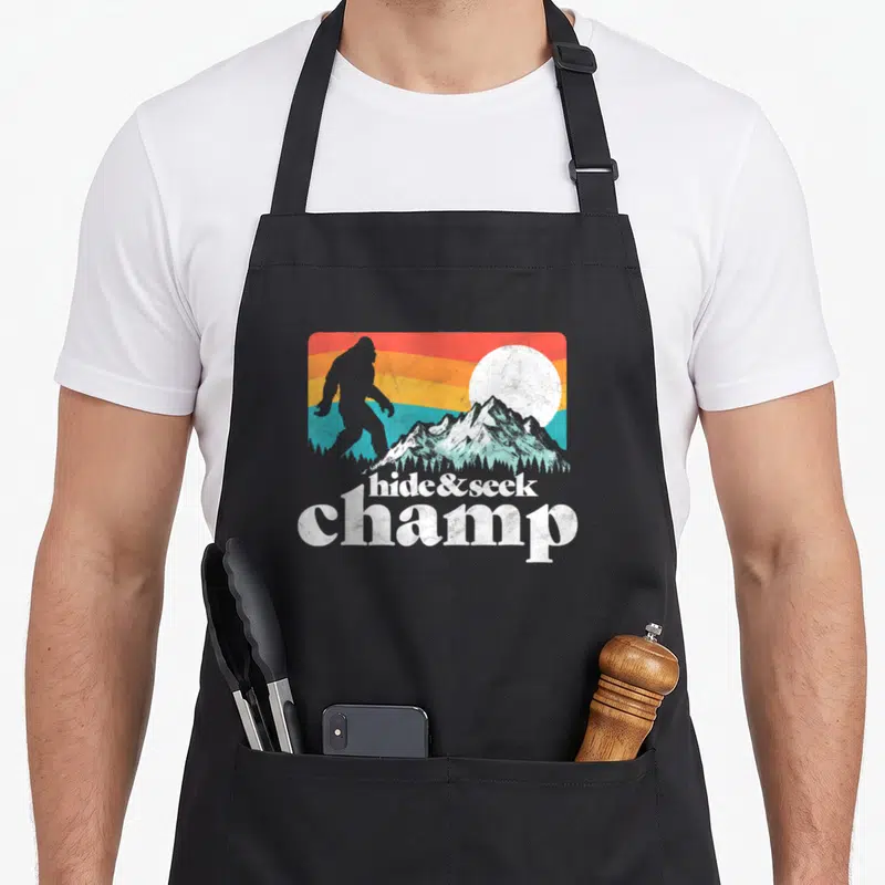 Champ bigfoot apron - Distressed hide and seek champ text below Bigfoot silhouette against colorful mountain sunset — shown on a black cotton canvas apron against a white background
