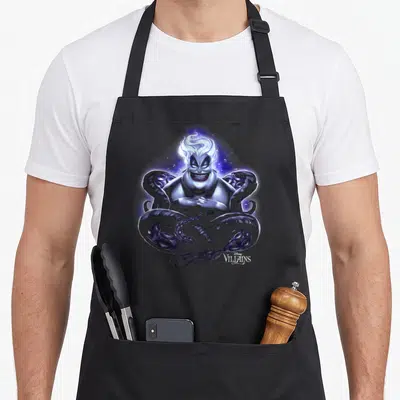 Ursula novelty apron - Ursula sea witch with swirling tentacles and Disney Villains logo in purple blue white cartoon — shown on a black cotton canvas apron against a white background