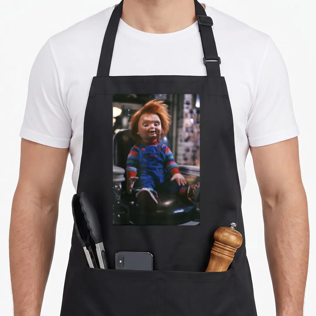 Sitting chair apron - Photo of Chucky doll in blue overalls sitting on black barber chair — shown on a black cotton canvas apron against a white background