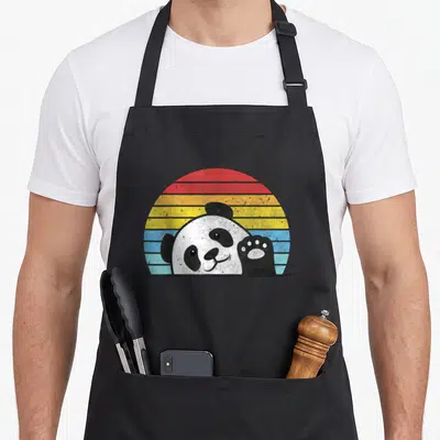 Zookeeper bear apron - Cartoon panda head and paw peeking over edge with red yellow blue striped sun — shown on a black cotton canvas apron against a white background
