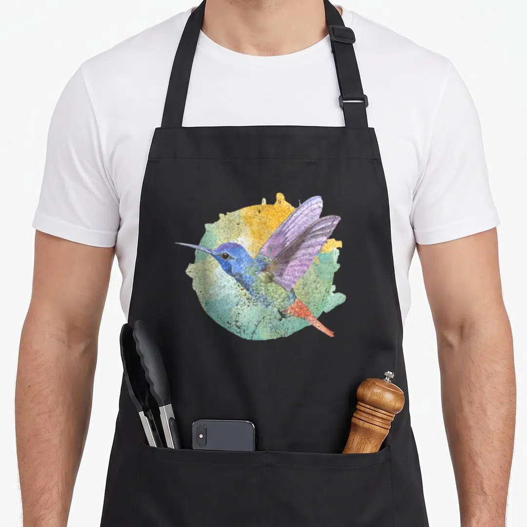 Colorful birds apron - Watercolor hummingbird with blue, purple, green, yellow, and orange circular background — shown on a black cotton canvas apron against a white background