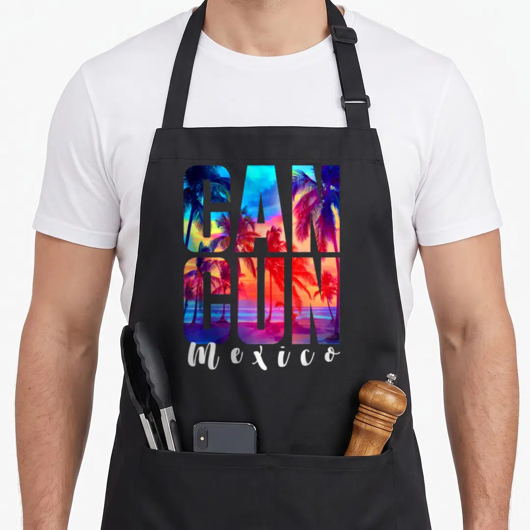 Palm mexico apron - CANCUN text filled with palm tree sunset in blue red yellow white purple and script Mexico below — shown on a black cotton canvas apron against a white background