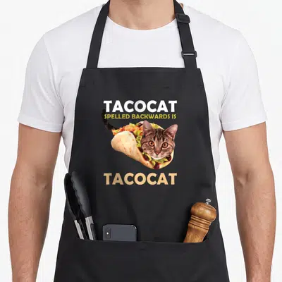 Tacocat cat taco apron - Cat head inside taco shell with TACOCAT text above and below — shown on a black cotton canvas apron against a white background