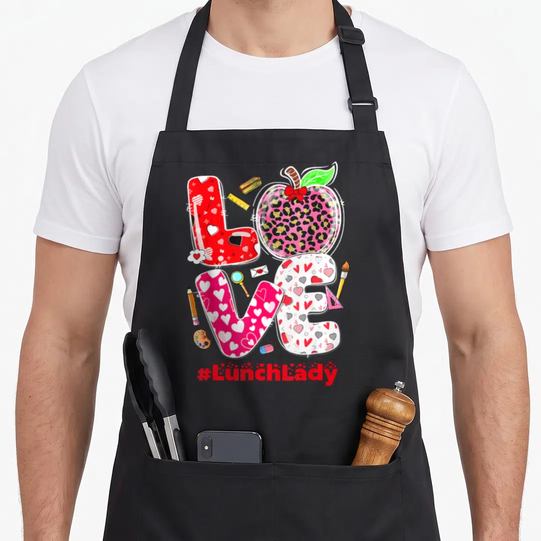 Valentine cafeteria aprons - LOVE text with leopard apple O, scattered school supplies, and #LunchLady hashtag in red — shown on a black cotton canvas apron against a white background