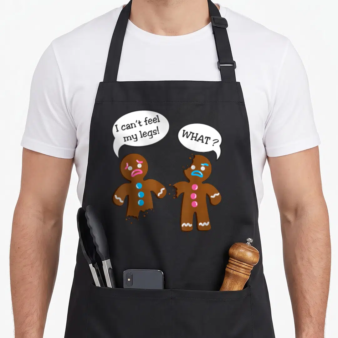 Christmas gingerbread apron - Two gingerbread men, one missing legs, with speech bubbles reading I can't feel my legs — shown on a black cotton canvas apron against a white background