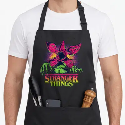 Season video game apron - Pixel art monster flower with green claws and STRANGER THINGS text in pink and yellow — shown on a black cotton canvas apron against a white background