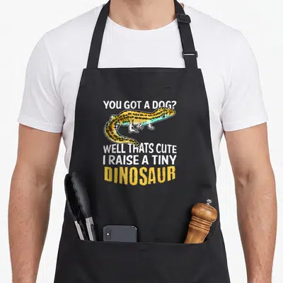 Pet leopard apron - Cartoon gecko in gold blue and yellow surrounded by white and yellow dinosaur joke text — shown on a black cotton canvas apron against a white background