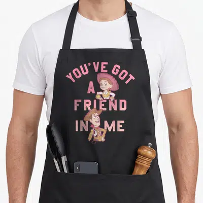 Friend dog breed aprons - Stacked pink text YOU'VE GOT A FRIEND IN ME with cartoon cowboy and cowgirl characters — shown on a black cotton canvas apron against a white background