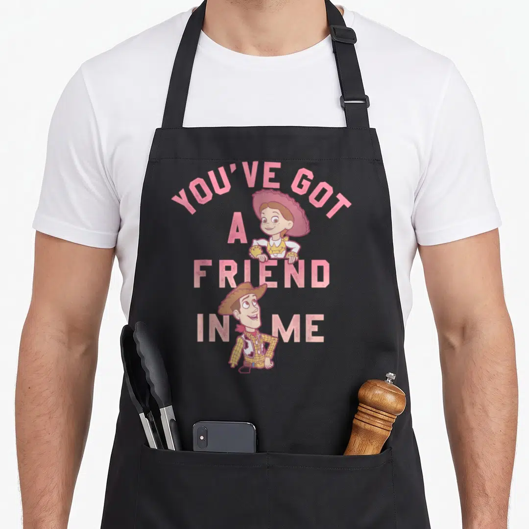Friend dog breed aprons - Stacked pink text YOU'VE GOT A FRIEND IN ME with cartoon cowboy and cowgirl characters — shown on a black cotton canvas apron against a white background