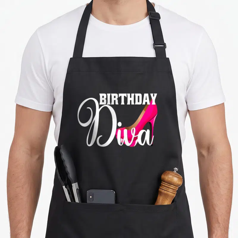 Birthday shoe apron - BIRTHDAY Diva text with pink high heel shoe overlapping white script on black background — shown on a black cotton canvas apron against a white background