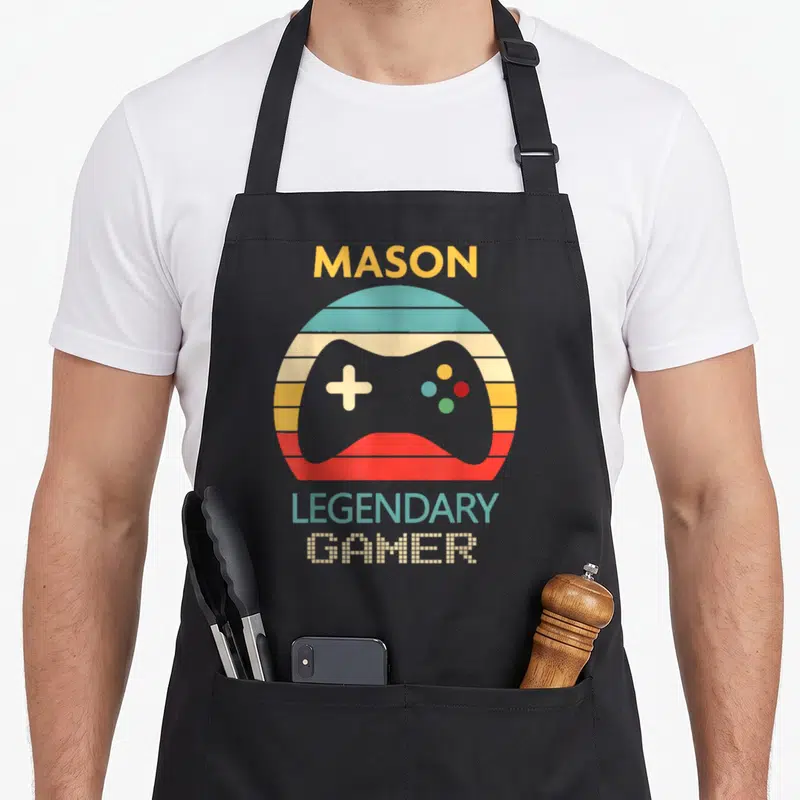 Legendary mason apron - Grey game controller over multi-colored striped circle with MASON and LEGENDARY GAMER text — shown on a black cotton canvas apron against a white background