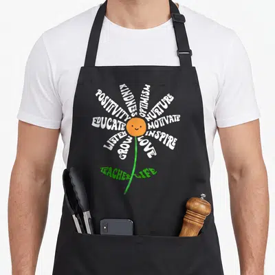 Inspirational teacher apron - Flower graphic with white word petals and orange smiley center reading teacher life — shown on a black cotton canvas apron against a white background