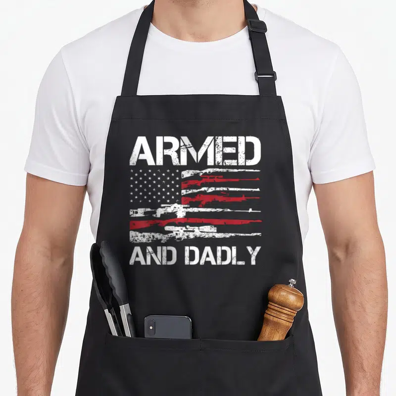 Dadly father day apron - Distressed USA flag with rifle stripes and text ARMED AND DADLY in white, red, and blue — shown on a black cotton canvas apron against a white background