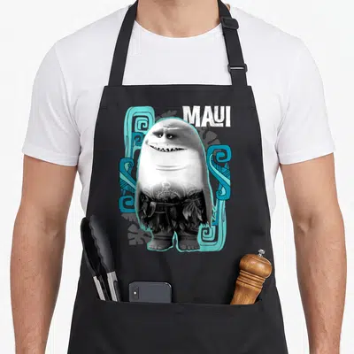 Shark novelty apron - Cartoon half-shark character with tribal tattoos and leaf skirt surrounded by blue patterns — shown on a black cotton canvas apron against a white background